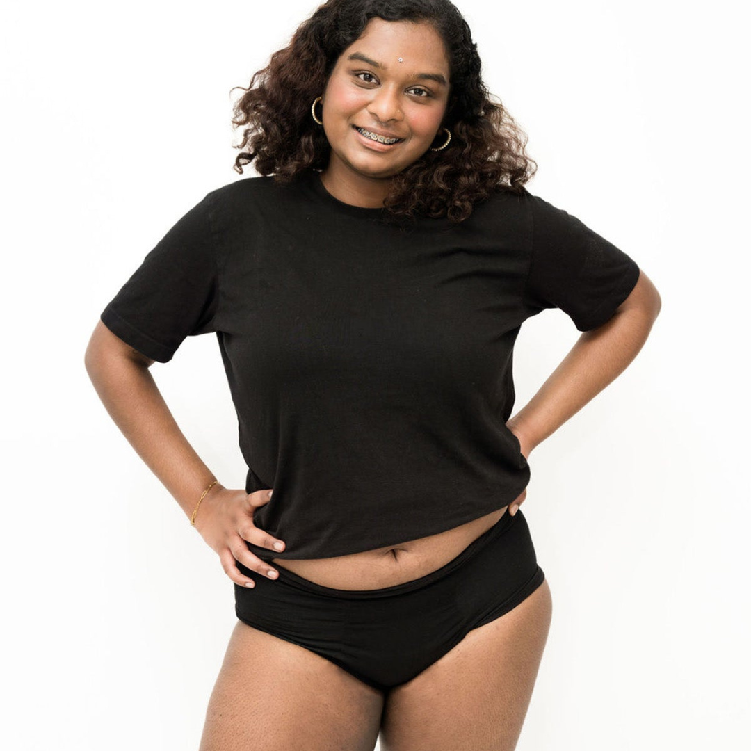 A lady wearing a black crewneck t-shirt and the Women’s Absorbent Underwear Midi in black, smiling with hands on hips against a white background.
